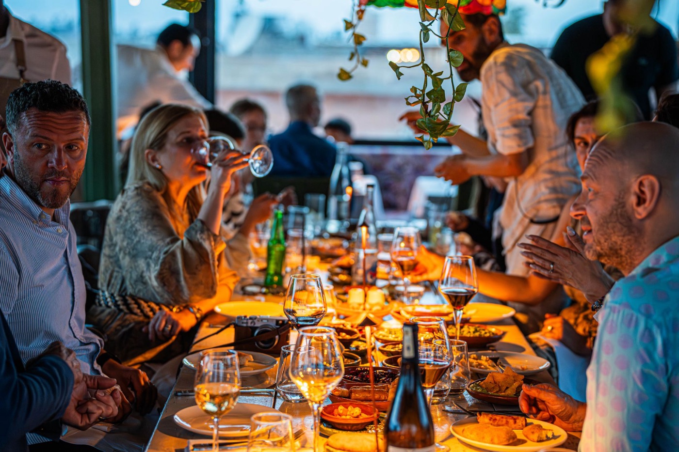 A long candlelit table of people talking over dinner